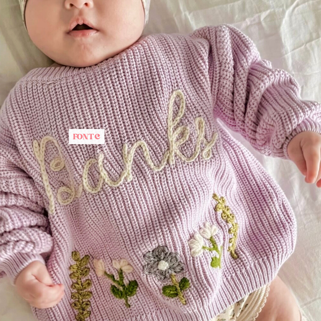 Baby girl in a purple knit sweater with flowers and a chain stitch name.